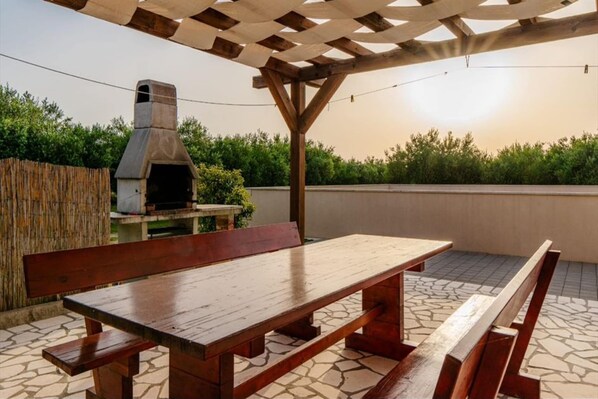 An outdoor patio with a wooden table and benches, a stone-tiled floor, and a pergola overhead.