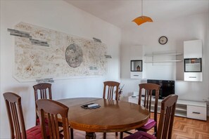 A dining area with a round wooden table, six wooden chairs, a large abstract painting on the wall, a clock, and a TV on a stand.