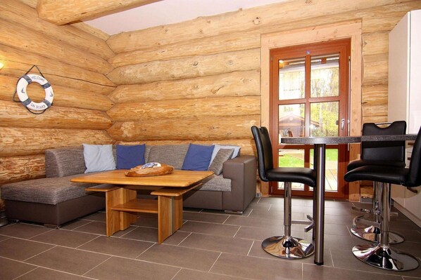 Interior de una cabaña de troncos con una mesa de madera, dos taburetes, un sofá con cojines y una puerta que da al exterior.