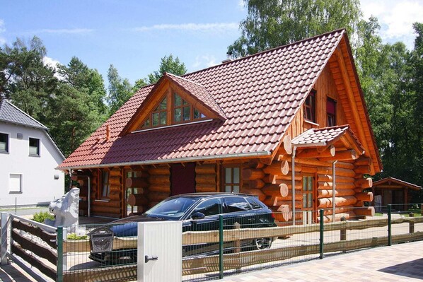 A log cabin with a red-tiled roof, a car parked in front, and a fenced area.