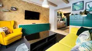 A modern living room with a yellow sofa, a glass coffee table, and a TV mounted on a wooden wall. There is a kitchen visible in the background with teal cabinets.