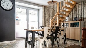 A dining area with a wooden table and chairs, a clock, a staircase, and a kitchen area with cabinets and appliances.