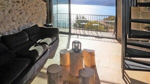 A modern living room with a black sofa, a stone wall, and a view of the sea through large glass doors. - Scenic Village Home with Lake Panorama (Sainte-Croix-du-Verdon)