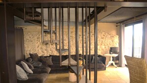 A modern living room with a stone wall, a dark-colored sofa, a smaller armchair, a coffee table, and a staircase with a metal railing. - Scenic Village Home with Lake Panorama (Sainte-Croix-du-Verdon)