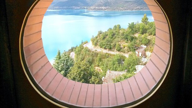 A circular window with a view of a lake, surrounding greenery, and distant mountains. - Scenic Village Home with Lake Panorama (Sainte-Croix-du-Verdon)