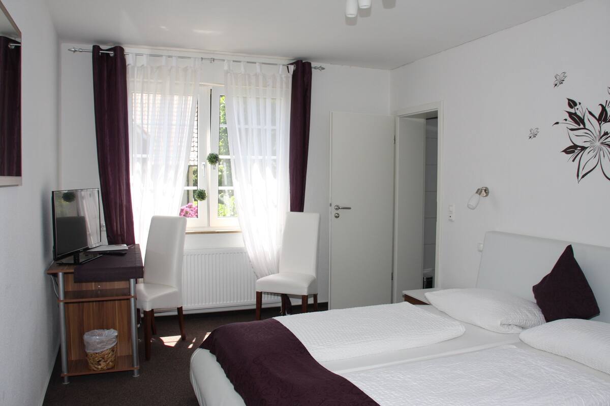 A hotel room with a bed, a desk with a computer, a chair, and a window with curtains.