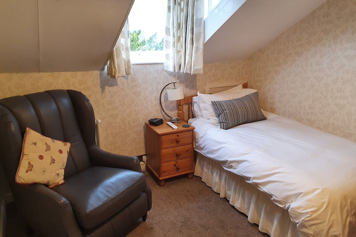 A bedroom with a single bed, a nightstand, a lamp, a chair, and a window with curtains.