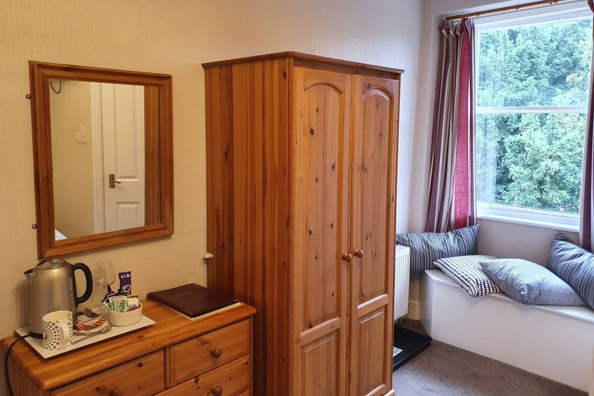 A room with a wooden wardrobe, a dresser with a mirror, and a window with a view of trees.