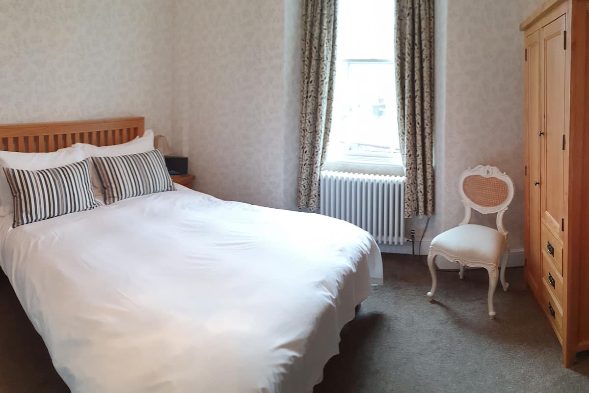 A bedroom with a large bed, a wooden nightstand, a chair, a wooden wardrobe, and a radiator.