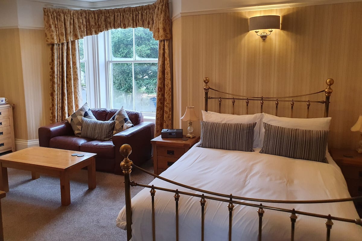A bedroom with a bed, a wooden nightstand, a leather sofa, a wooden table, and a window with curtains.