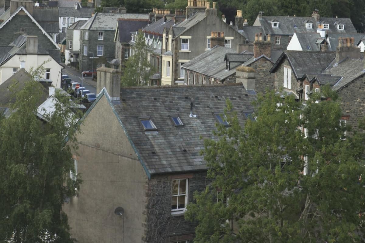 A town with traditional stone buildings, a street with parked cars, and a backdrop of green hills.