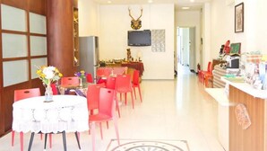 Un hall de hotel con una recepción, un televisor montado en la pared y un comedor con sillas rojas y una mesa blanca.