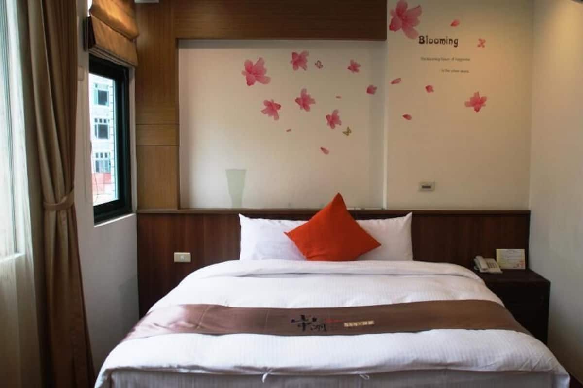 A hotel room with a bed, a nightstand, a window with curtains, and a wall with a floral decal.