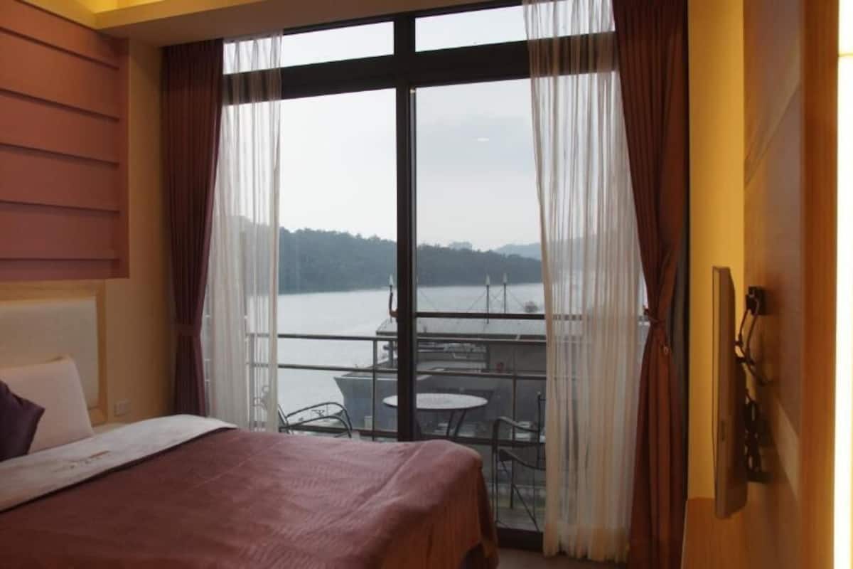 A hotel room with a bed, a view of a body of water, and a balcony with a table and chairs.