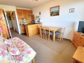 Een gezellige kamer met een houten bank, een eettafel met stoelen, een keuken met houten kasten en een televisie op een houten dressoir.