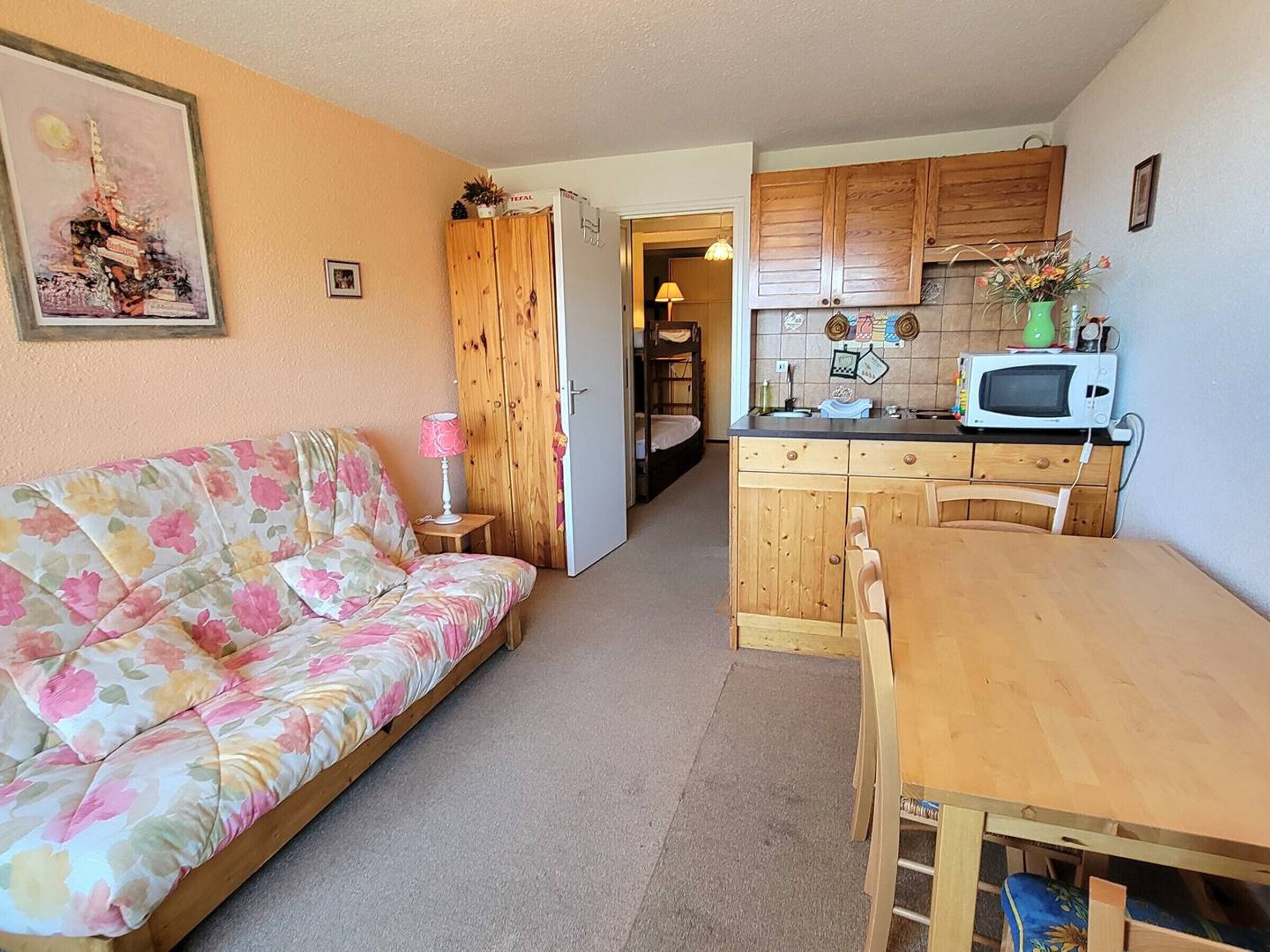 A cozy living room with a floral sofa, a wooden dining table, and a kitchen area with wooden cabinets and a microwave.