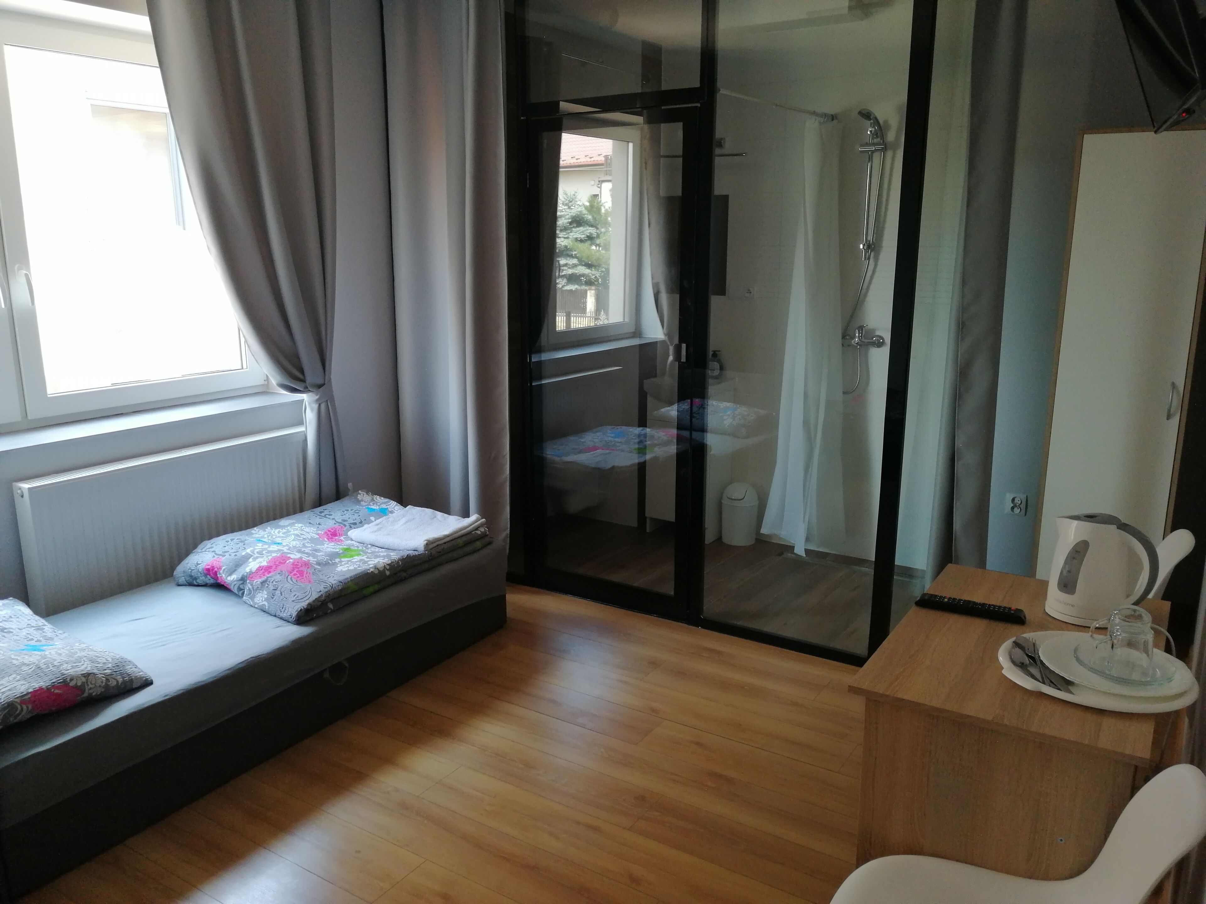 A room with a bed, a small table with a kettle, a chair, and a glass shower enclosure.
