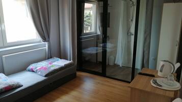 A room with a bed, a small table with a kettle, a chair, and a glass shower enclosure.