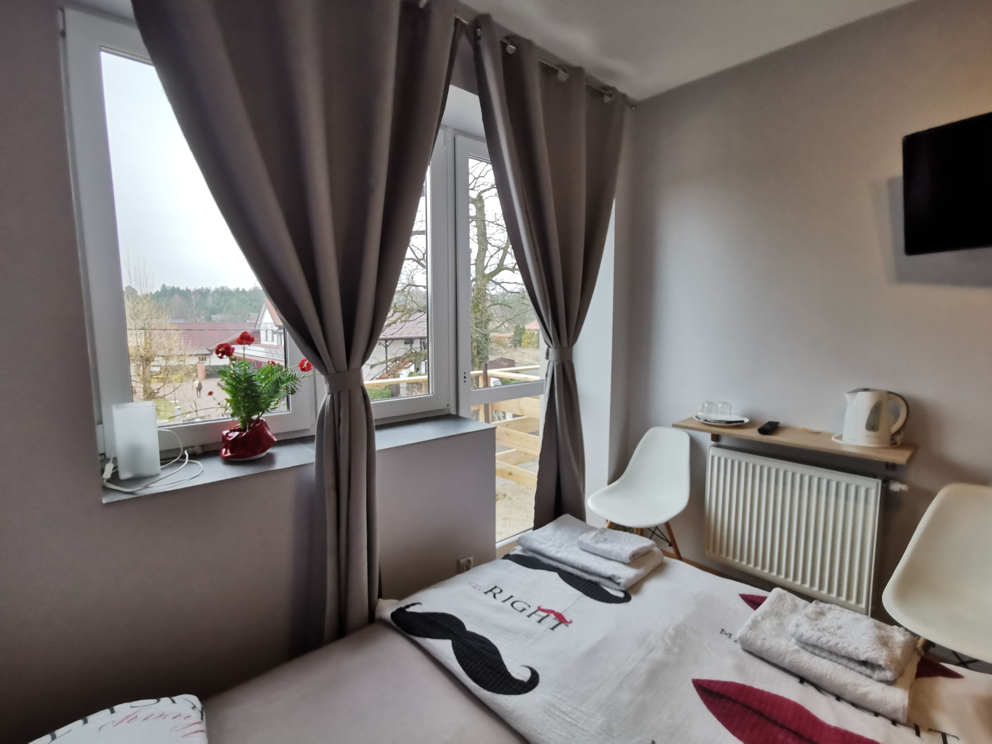 A bedroom with a bed, a window with curtains, a radiator, a chair, and a small table with a kettle and a cup.