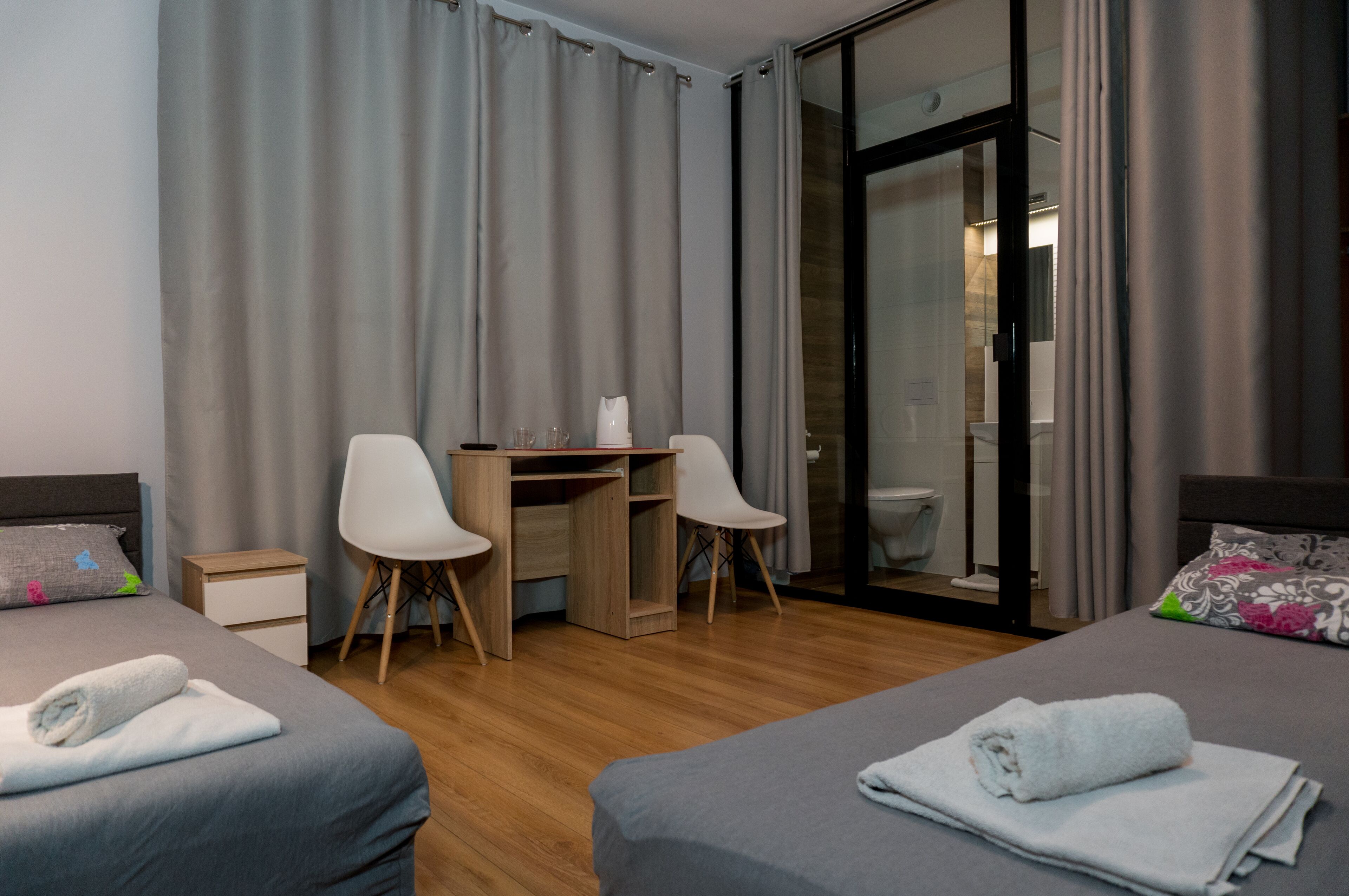 A hotel room with two beds, a desk, two chairs, a small bedside table, and a bathroom visible through a glass door.