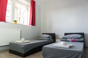 Quarto com duas camas, janela com cortinas vermelhas, radiador e uma pequena planta no parapeito.