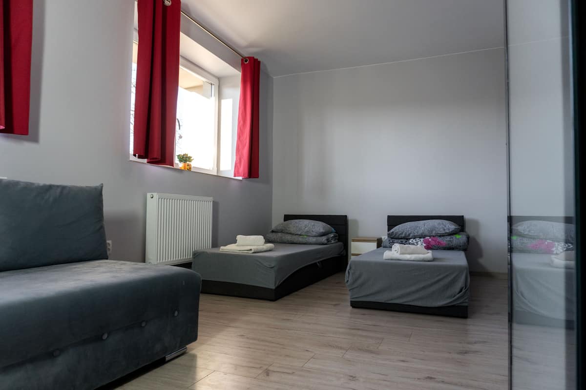 Une chambre moderne avec deux lits simples, un canapé gris et une fenêtre avec des rideaux rouges.