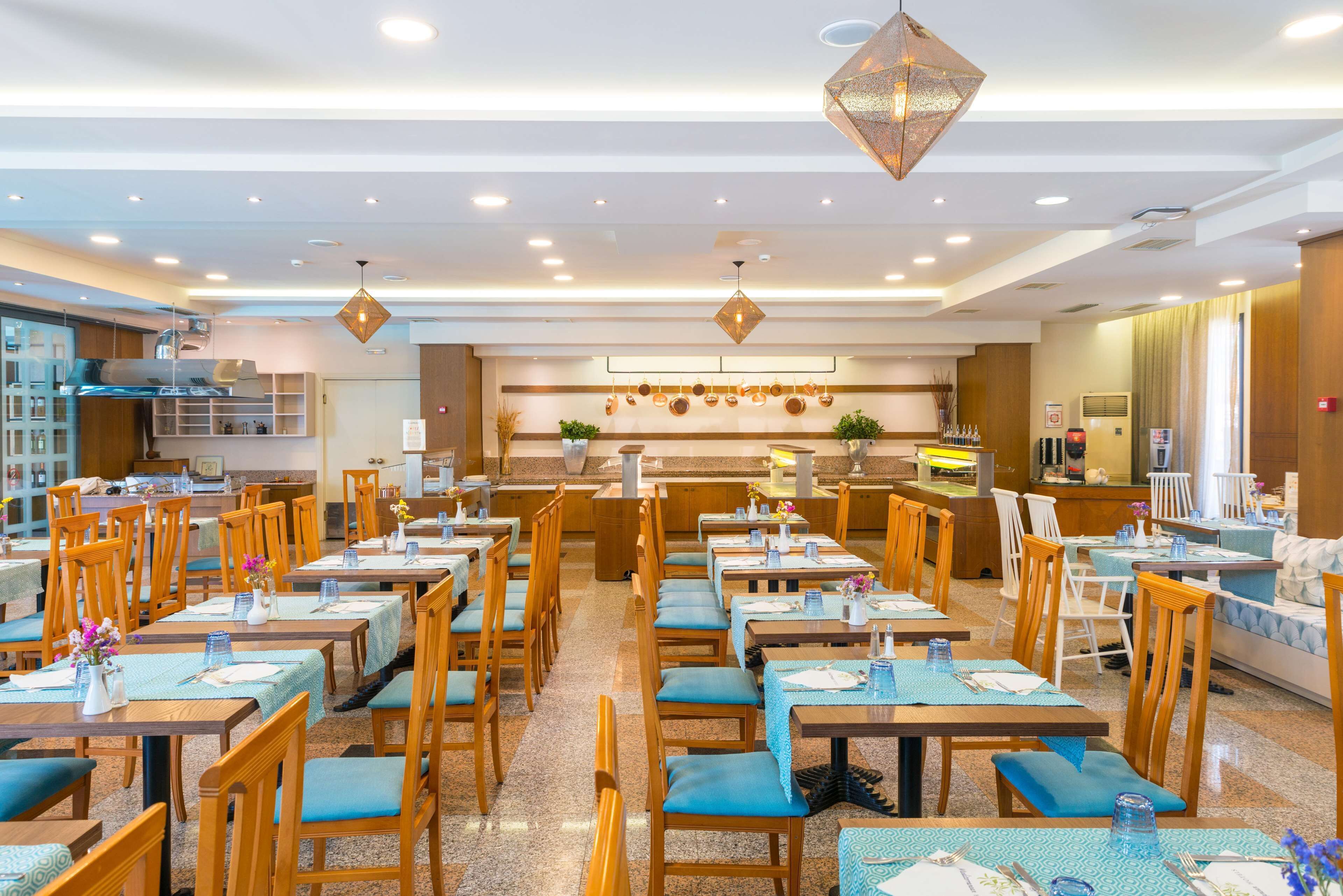 A spacious restaurant with multiple dining tables and chairs, a kitchen area in the background, and pendant lighting.