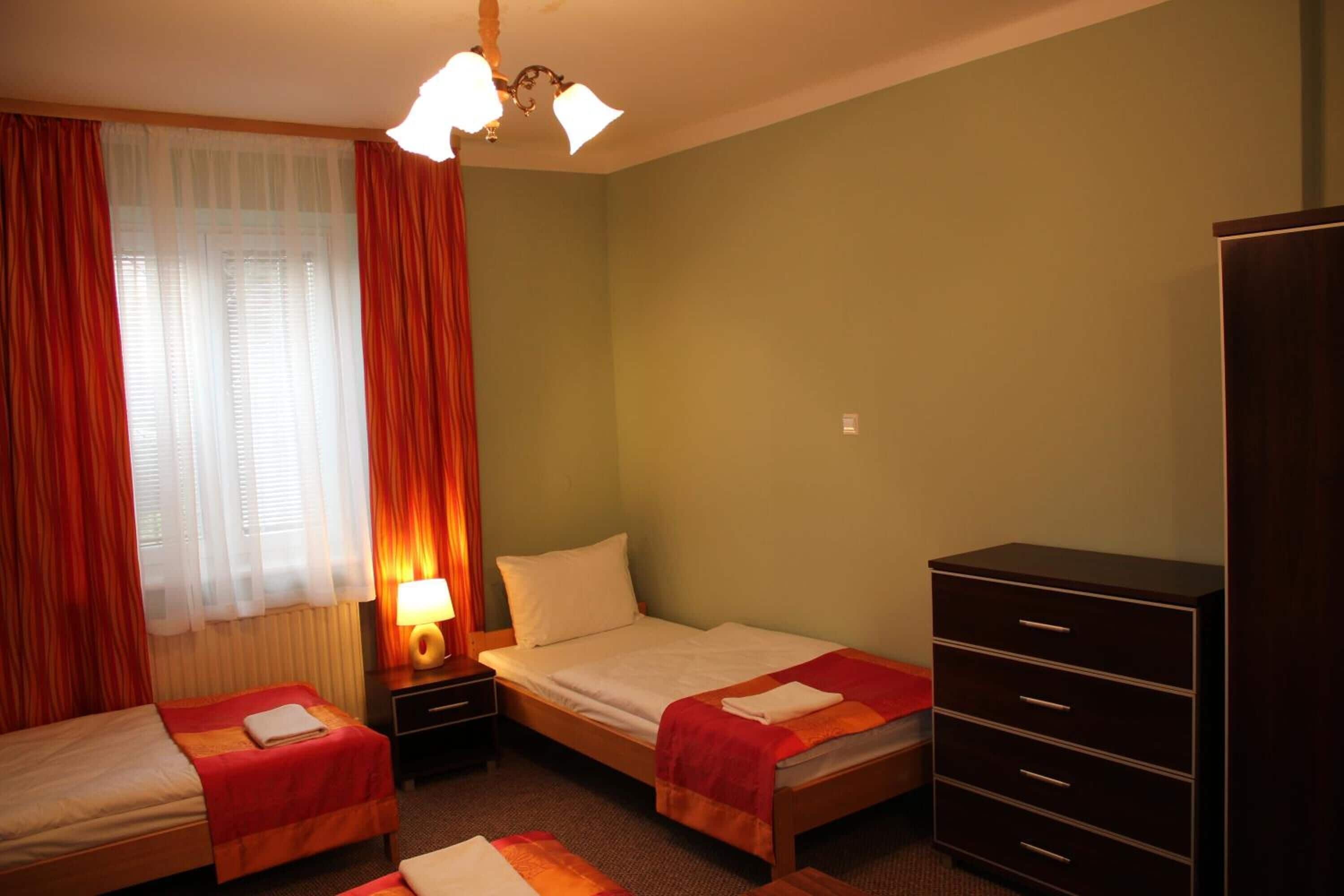 A hotel room with two single beds, a nightstand with a lamp, a dresser, and a window with curtains.