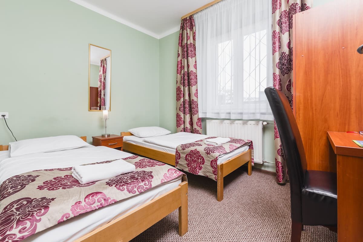A hotel room with two single beds, a wooden desk, a chair, a mirror, and a window with curtains.