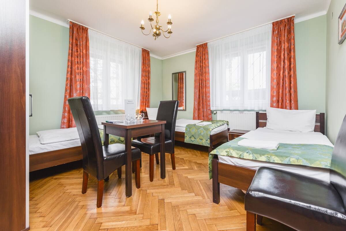 A room with two beds, a table with chairs, a chandelier, and large windows with curtains.