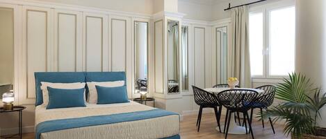 A bedroom with a bed, bedside tables, a dining area with chairs, a window with curtains, and a potted plant.