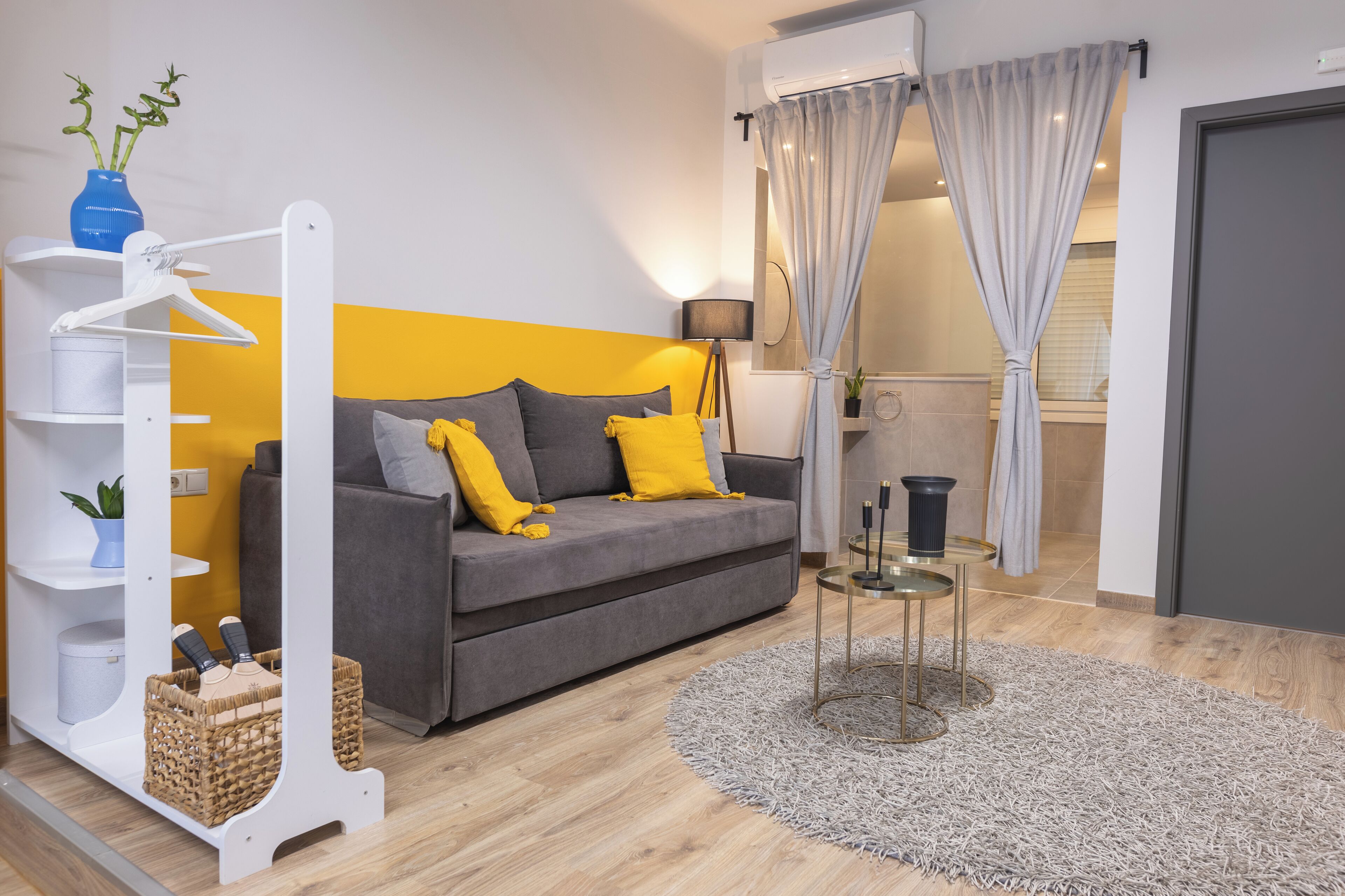 A modern living room with a grey sofa, yellow cushions, a white shelving unit, a small round table, and a grey shag rug.