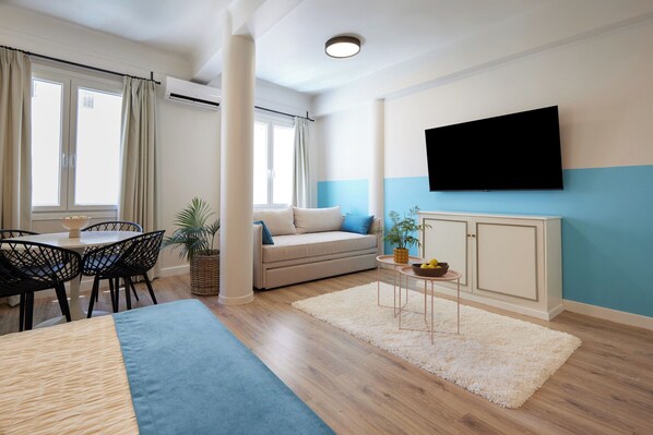 A modern living room with a sofa, a dining table with chairs, a TV mounted on the wall, and a large window with curtains.