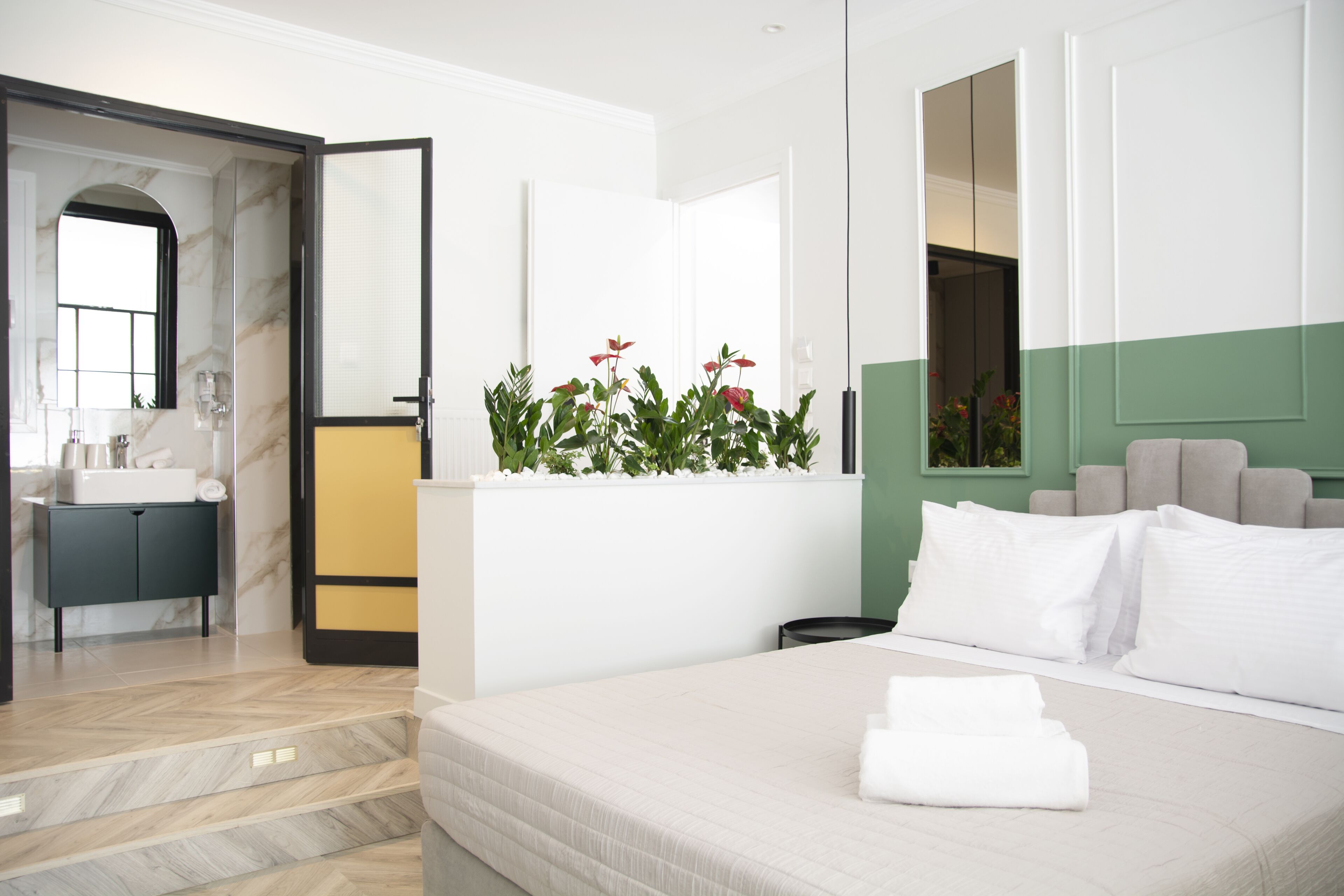A modern hotel room with a bed, a bedside table, a mirror, a plant, and a view of the hallway.