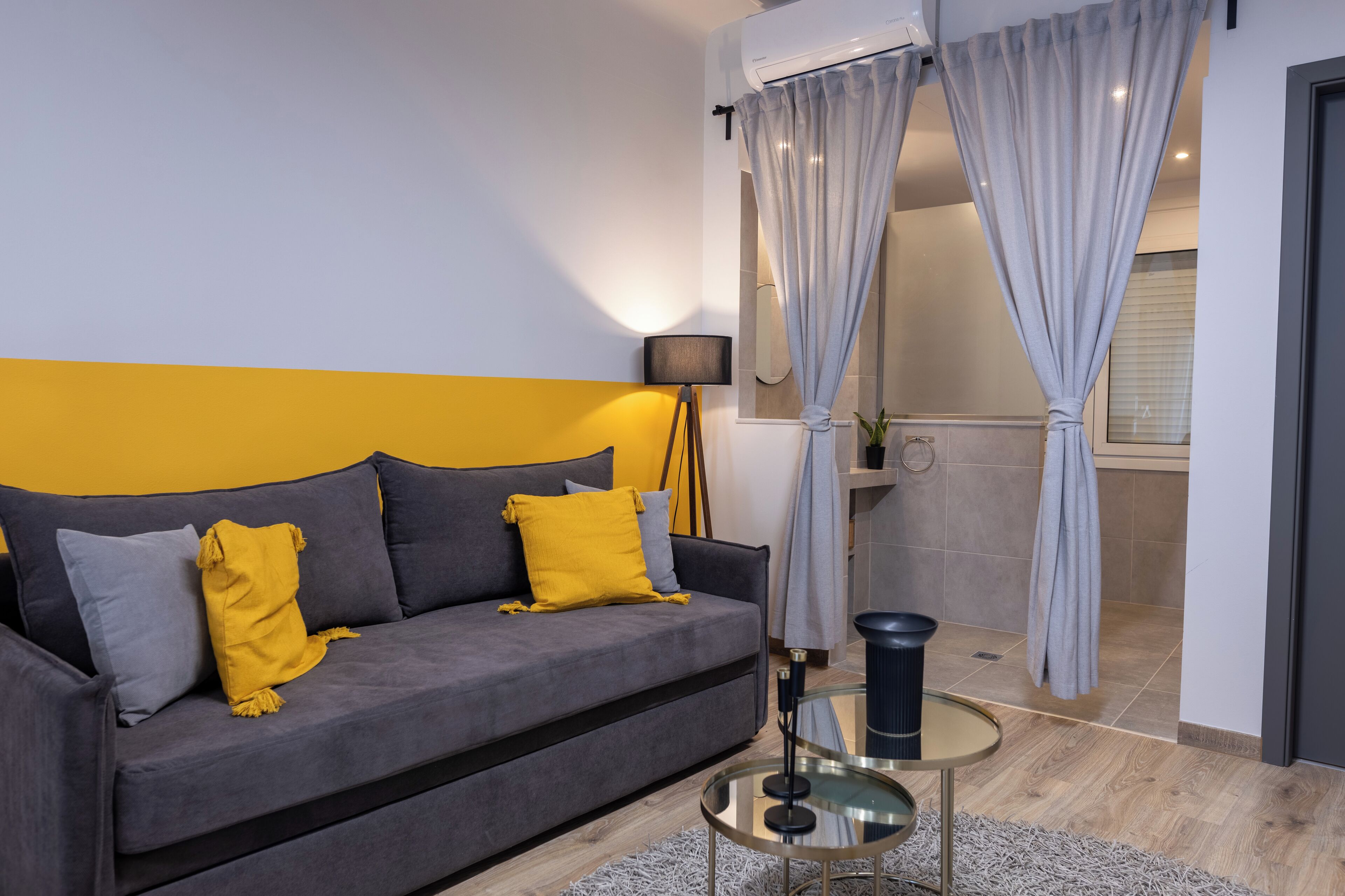 Una sala de estar moderna con un sofá gris, cojines amarillos y grises, una mesa de centro de vidrio y una cortina gris.