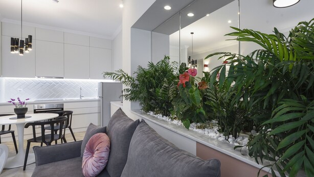 Un salon moderne avec un canapé gris, un coin repas avec une table et des chaises blanches, et une cuisine avec des meubles blancs et un dosseret en motif chevrons.