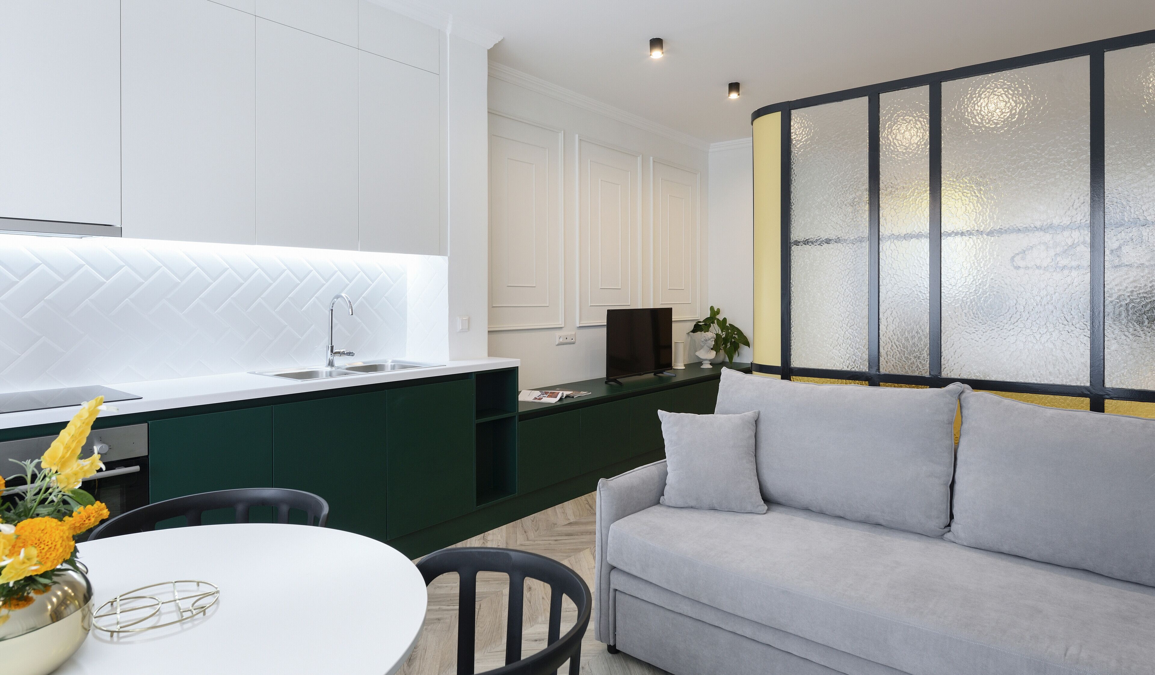A modern kitchen with a white backsplash, dark green cabinets, and a grey sofa in a living area with a large window.