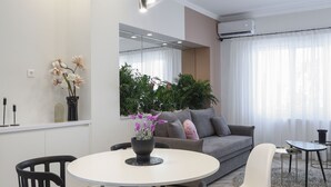 Un salon moderne avec une table à manger ronde, des chaises noires, un canapé gris et un fauteuil blanc. Une grande fenêtre est agrémentée de rideaux légers, un miroir est accroché au mur et une plante en pot est posée sur le rebord de la fenêtre.