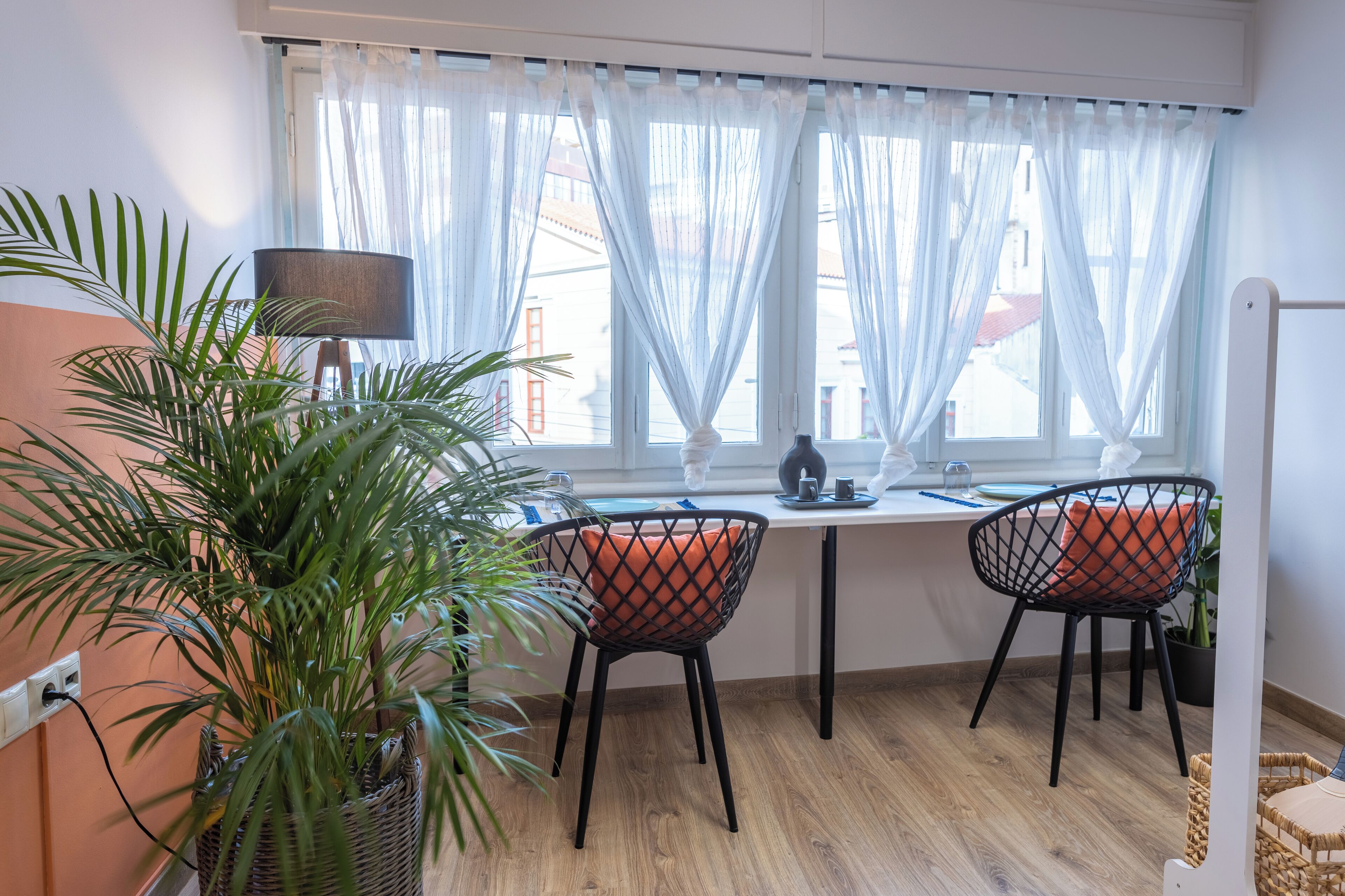 Quarto com mesa de jantar, duas cadeiras, planta em vaso, luminária e janela com cortinas translúcidas.