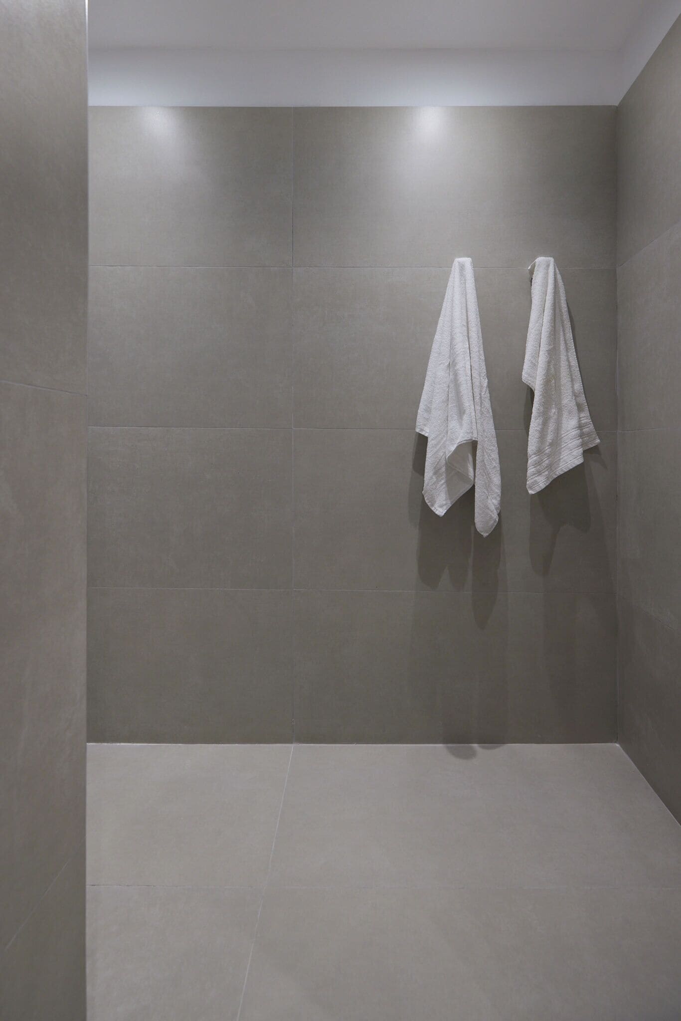 Un bagno moderno con piastrelle grigie, due asciugamani bianchi appesi a ganci e un design pulito e minimalista.