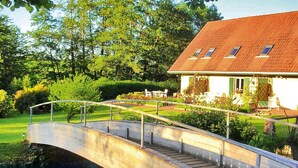 A concrete bridge over a body of water leading to a house with a red roof and white walls, surrounded by greenery. - Holiday Farm in Pfaffenhofen with Indoor Pool (Kindwiller)