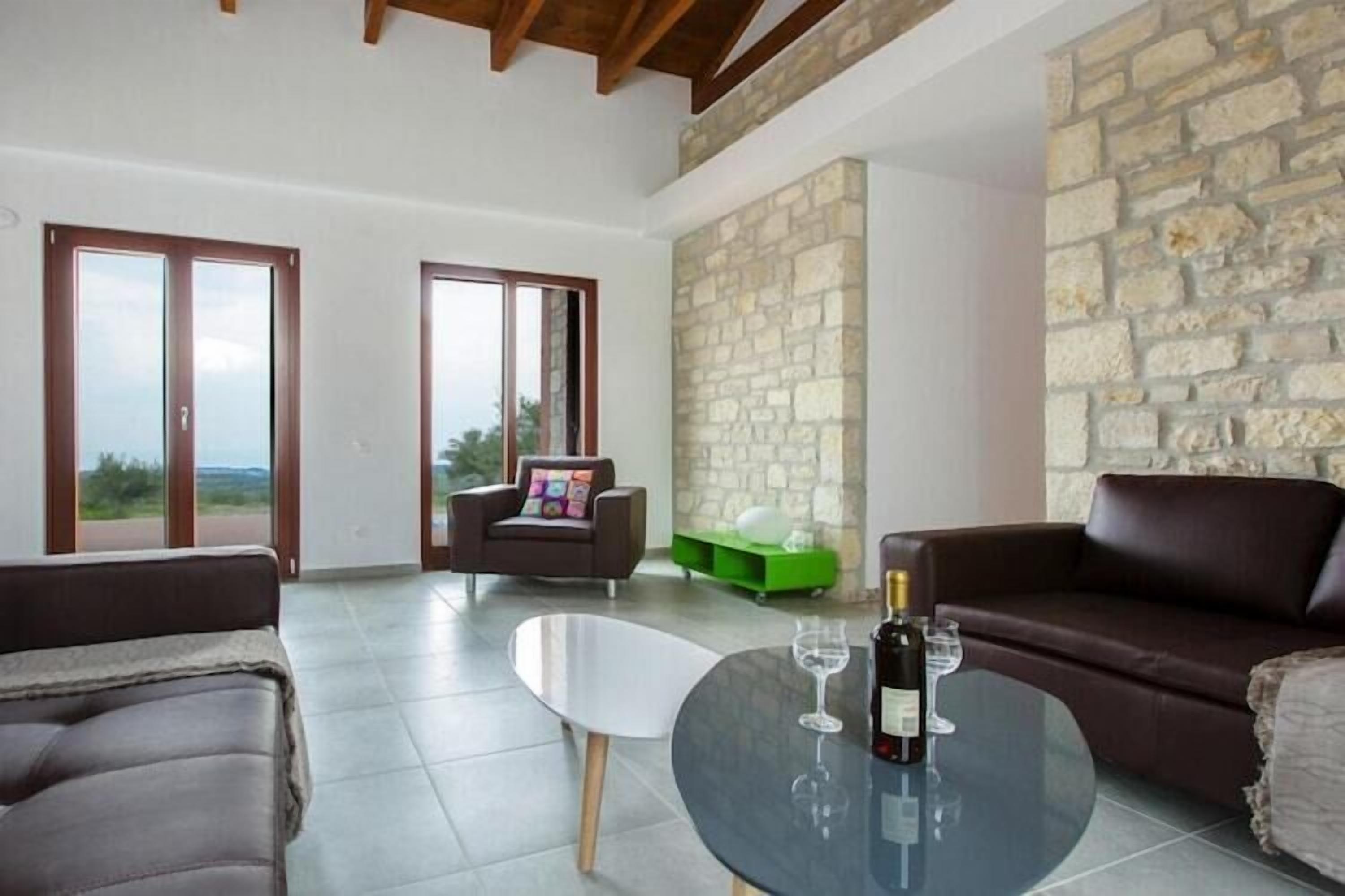 Amplia sala de estar con pared de piedra, sofás de cuero, una mesa de centro de vidrio y vistas a través de amplios ventanales correderas.