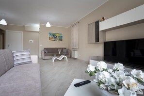 A modern living room with a sofa, a coffee table with a remote control, a TV, and a decorative flower arrangement. - Sicano Home (Delia)