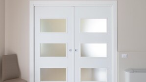 Una puerta blanca con paneles de vidrio esmerilado, una silla color beige y un termostato en la pared.