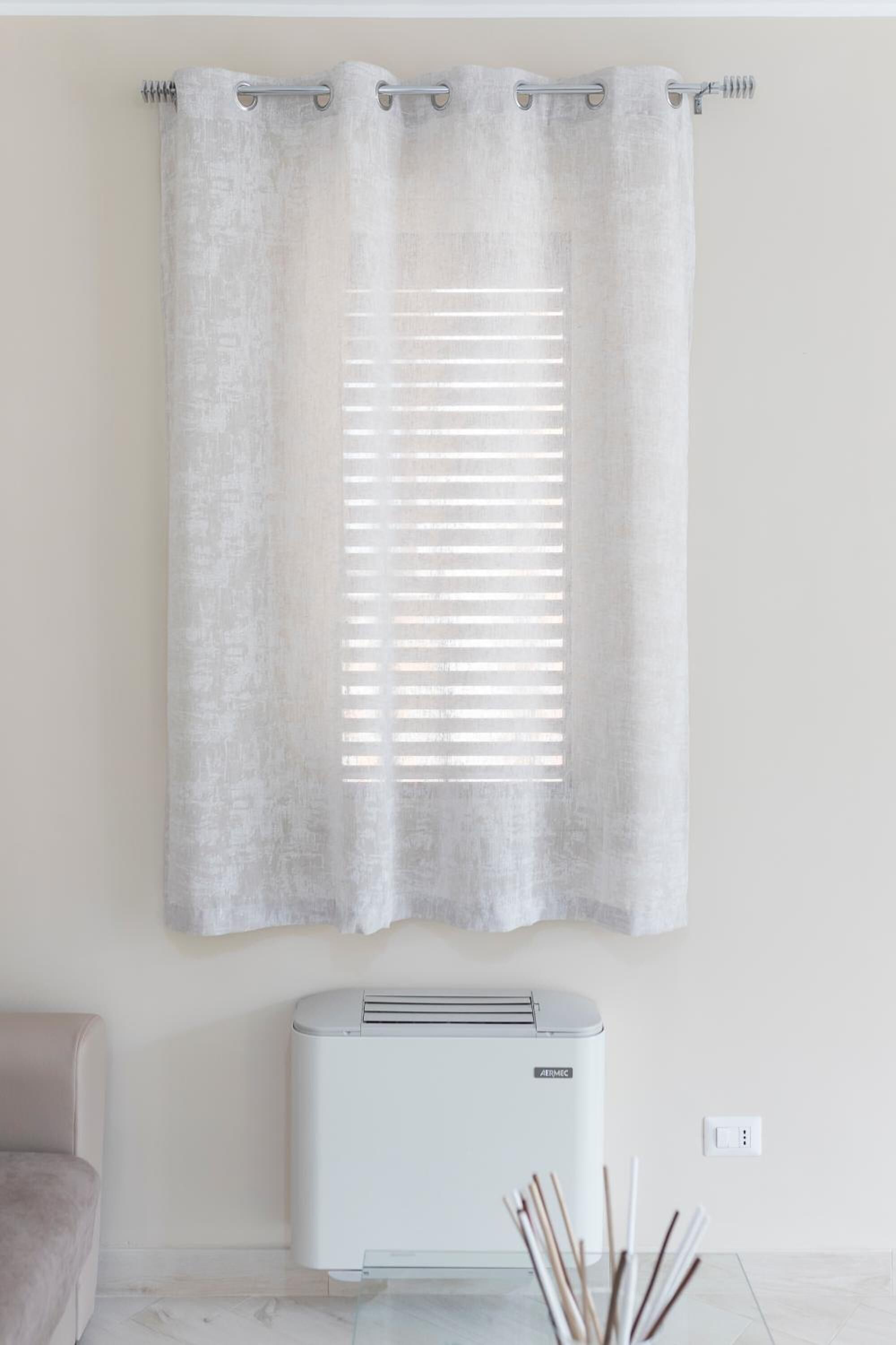 A room with a window covered by a light-colored curtain, a white air conditioning unit below the window, and a glass table with a few decorative items.