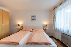 A double bed with two pillows, a wooden wardrobe, bedside lamps, a framed picture on the wall, and a window with sheer curtains.