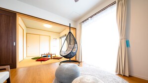 Uma sala de estar moderna com uma cadeira suspensa, dois pufes e vista para um espaço tradicional japonês ao fundo.