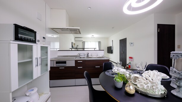 A modern kitchen with a built-in oven, microwave, and a large island with a sink. There is a dining area with a black table set for two, featuring a decorative centerpiece and a vase. - Erable Mt.Fuji Zen (Fujikawaguchiko)
