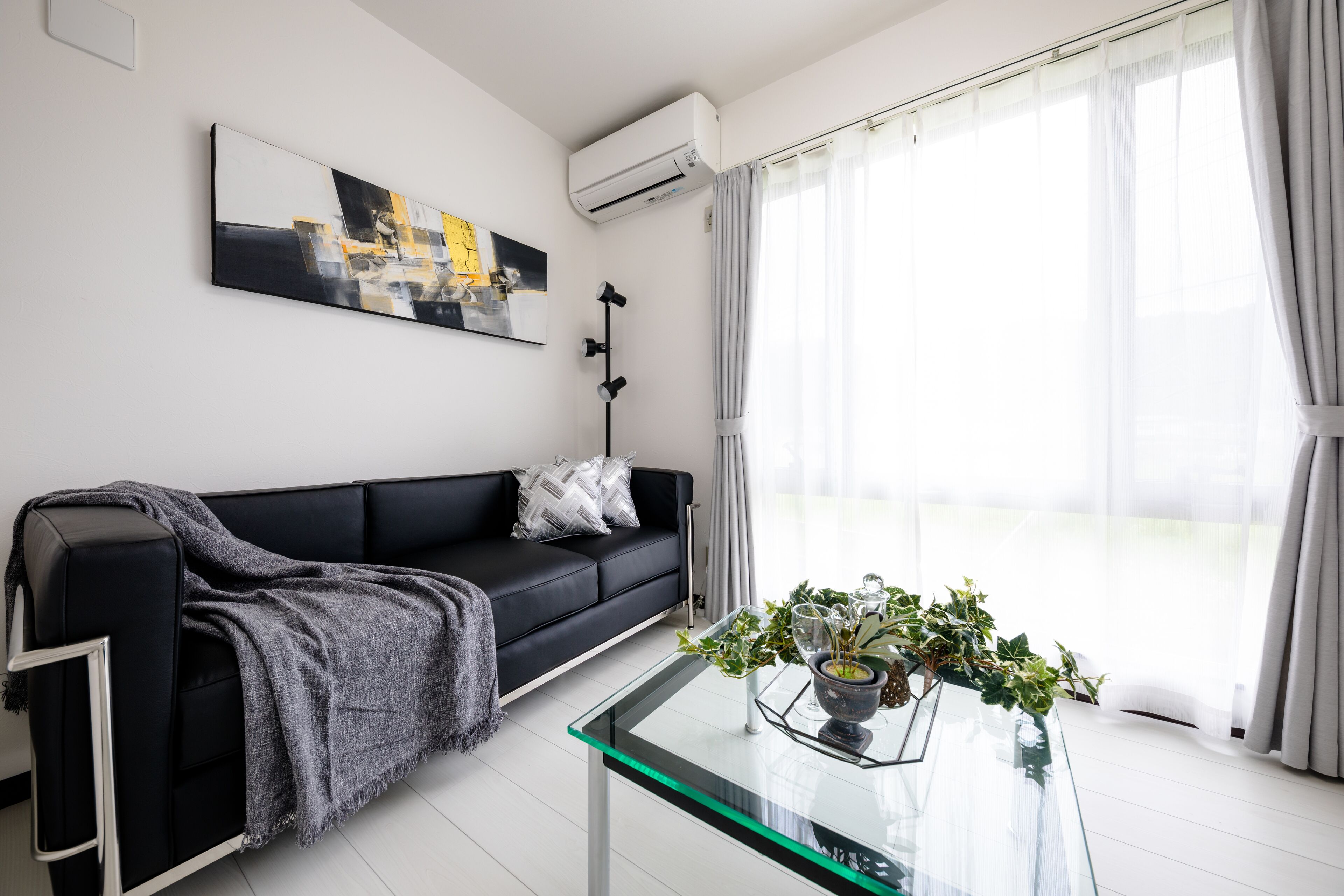 A modern living room with a black sofa, a glass coffee table with a plant, and a large window with curtains.