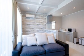 Uma sala de estar moderna com sofá em formato de L, uma área de cozinha com eletrodomésticos e uma parede com textura de pedra.
